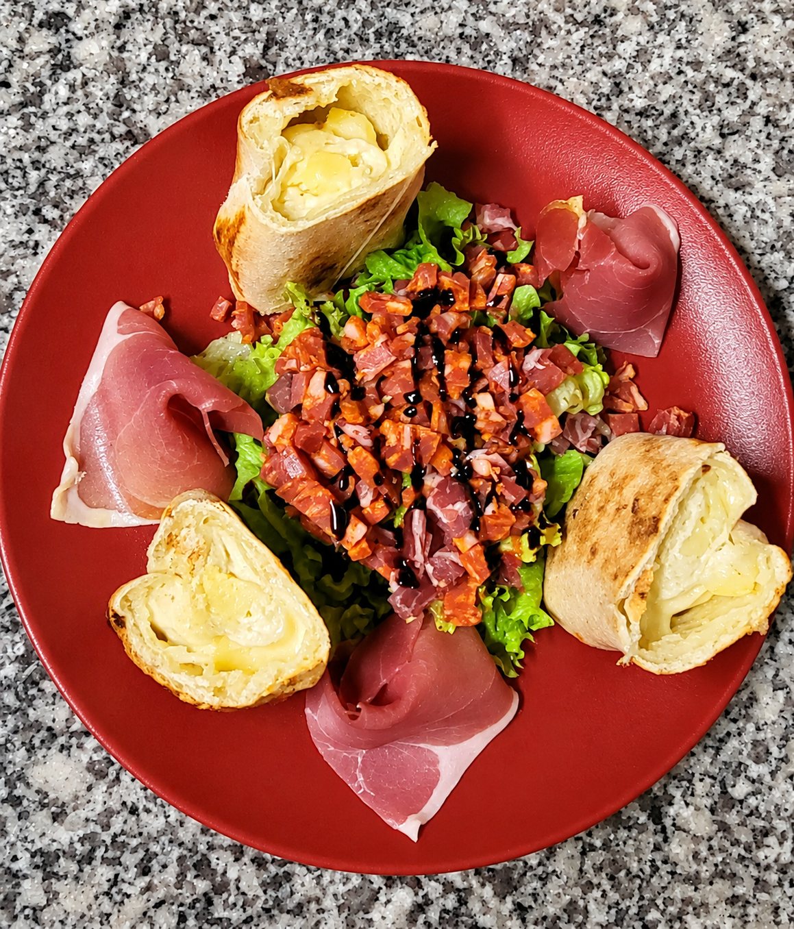 Vue de dessus d'une assiette rouge garnie de salade verte, de dés de chorizo avec glaçage balsamique, de tranches de jambon cru et de rouleaux feuilletés au fromage, posée sur un plan de travail en granit.