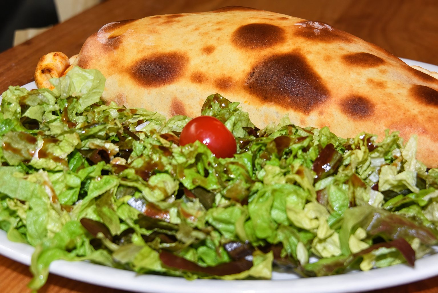 Une assiette blanche présente un calzone doré aux taches de cuisson brunes, accompagné d'une salade verte et d'une tomate cerise, posée sur une table en bois.
