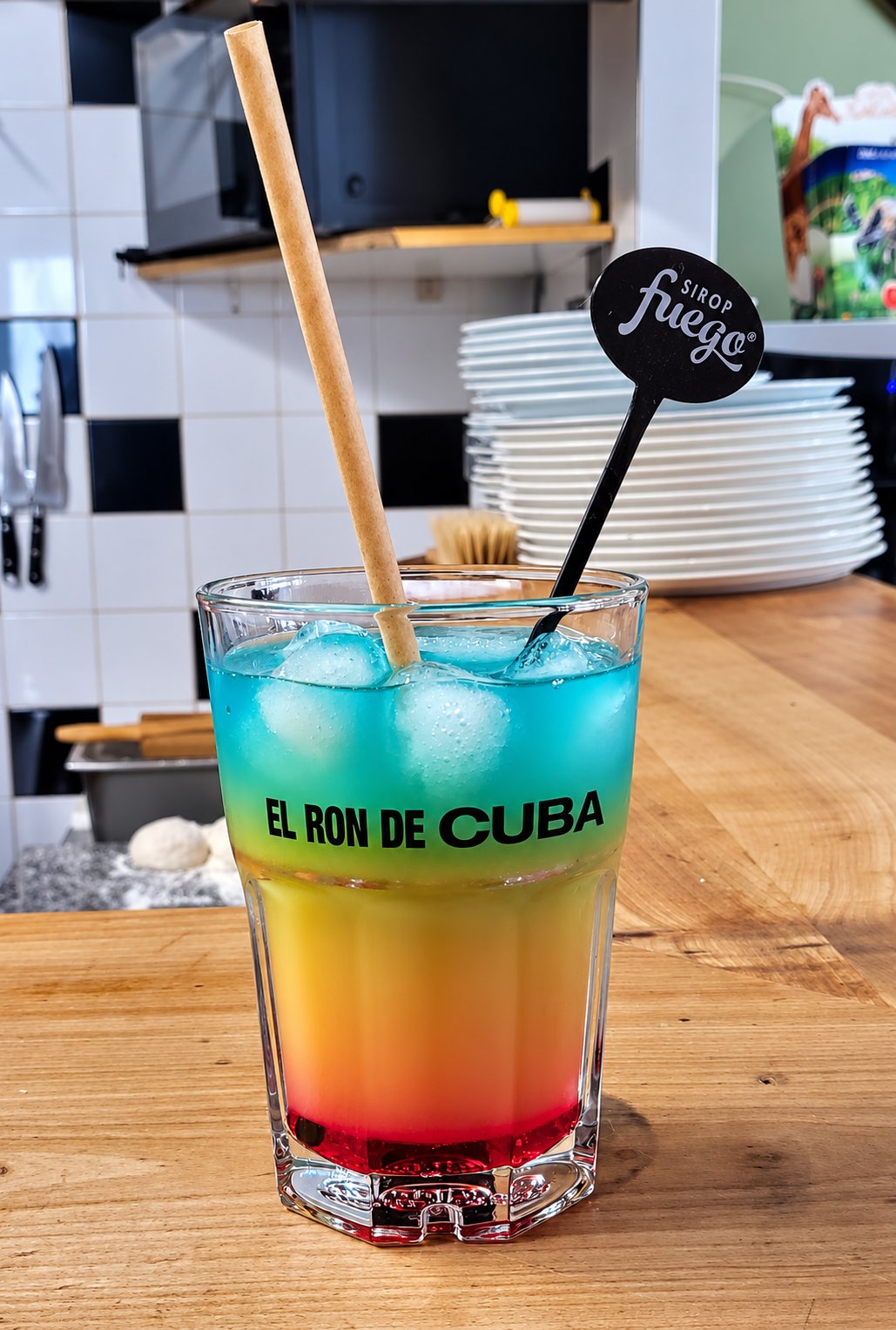 Un verre de cocktail en couches bleu, jaune et rouge avec des glaçons, une paille en papier et un agitateur noir Fuego, posé sur un comptoir en bois.