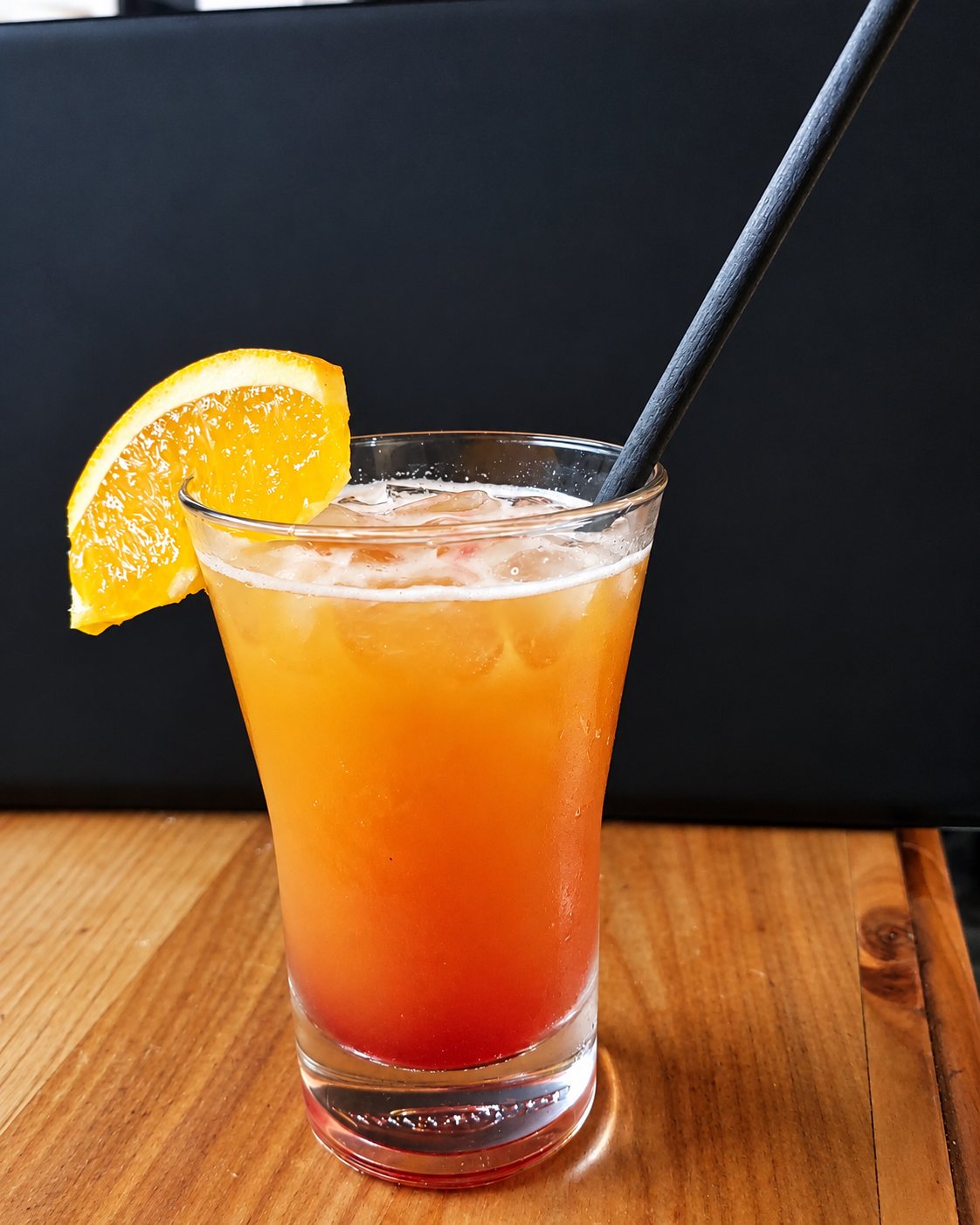 Un verre de cocktail dégradé orange et rouge avec des glaçons, une paille noire et une tranche d'orange, posé sur une table en bois.