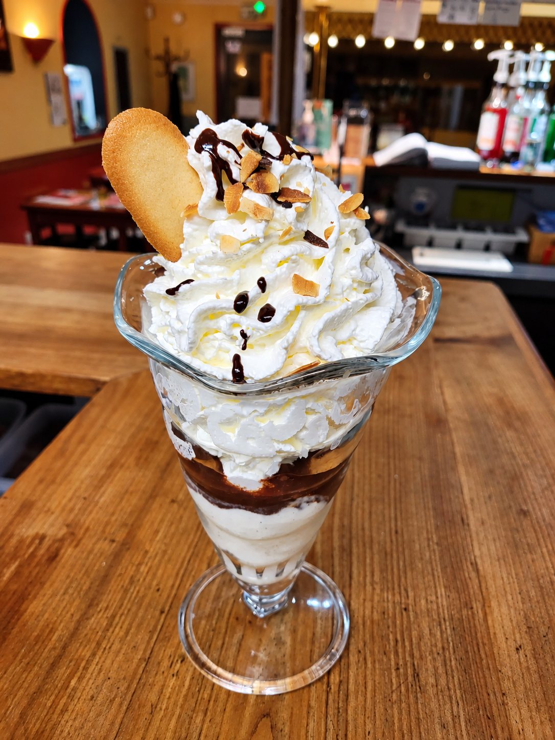 Une coupe glacée surmontée de chantilly, de sauce au chocolat, d'amandes effilées grillées et d'une tuile, posée sur un comptoir en bois dans un café.