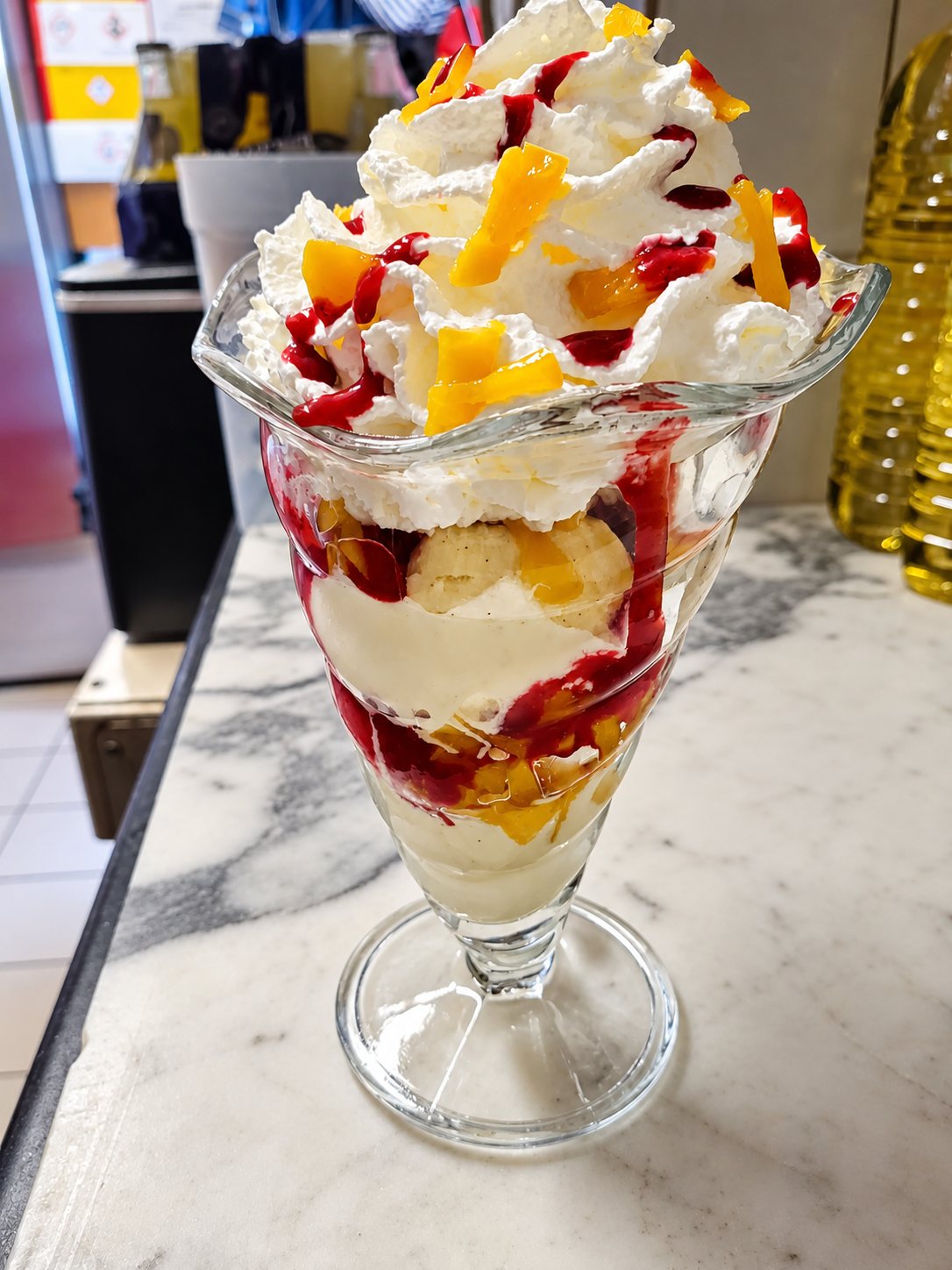 Une coupe glacée en verre surmontée de chantilly, de morceaux de mangue et de coulis rouge, posée sur un comptoir en marbre.