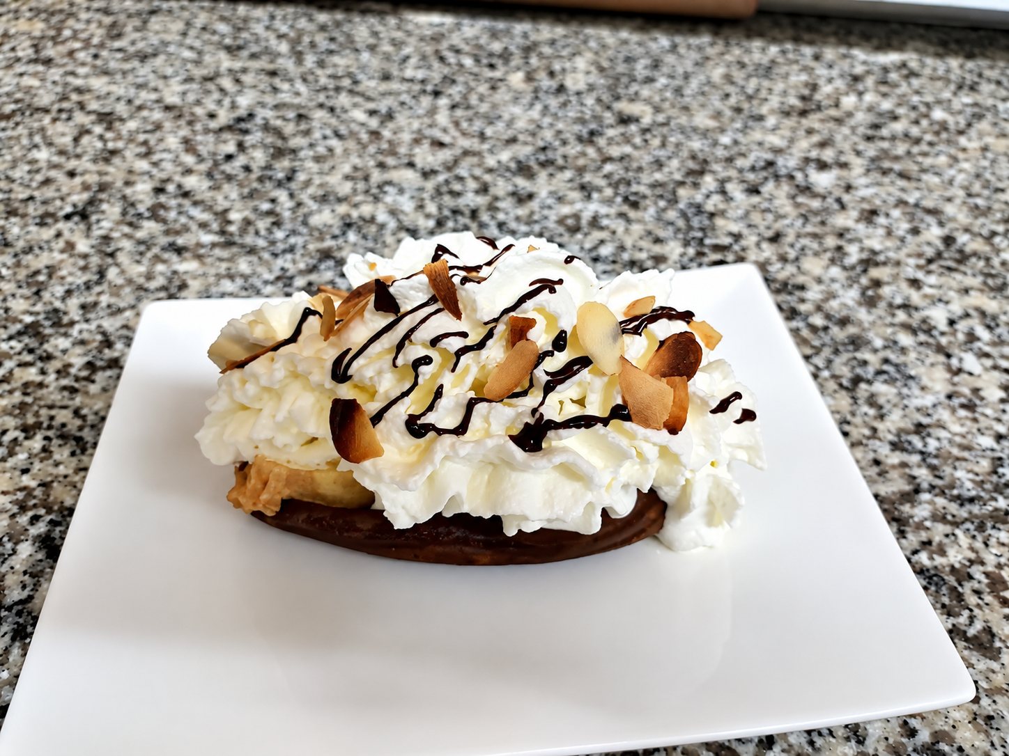 Un dessert garni de chantilly, d'amandes effilées grillées et de filets de chocolat, servi sur une assiette carrée blanche posée sur un plan de travail en granit.