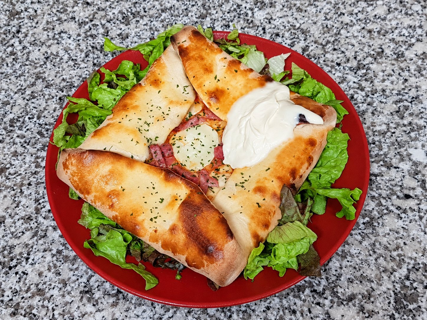 Une galette repliée et gratinée, garnie de fromage de chèvre, de charcuterie et de crème, servie sur un lit de salade dans une assiette rouge posée sur un plan de travail en granit.