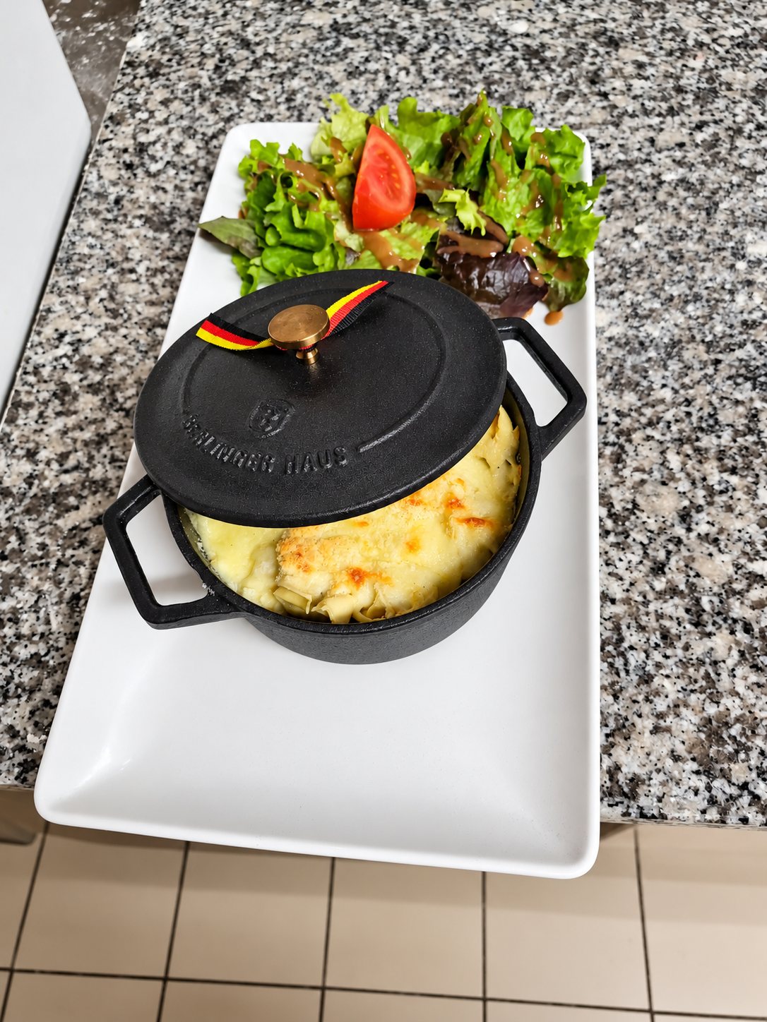Une petite cocotte en fonte noire contenant un gratin doré, posée sur un plateau blanc avec une salade verte et un quartier de tomate.