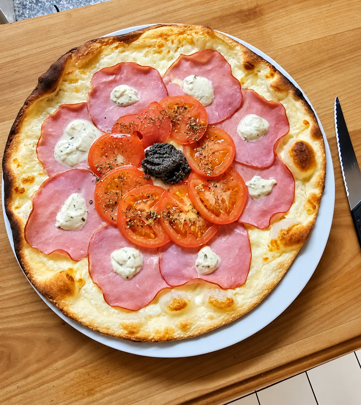 Pizza ronde sur une assiette blanche posée sur une table en bois, garnie de tranches de jambon rose, de rondelles de tomate, de fromage frais et d'une noix de tapenade au centre.