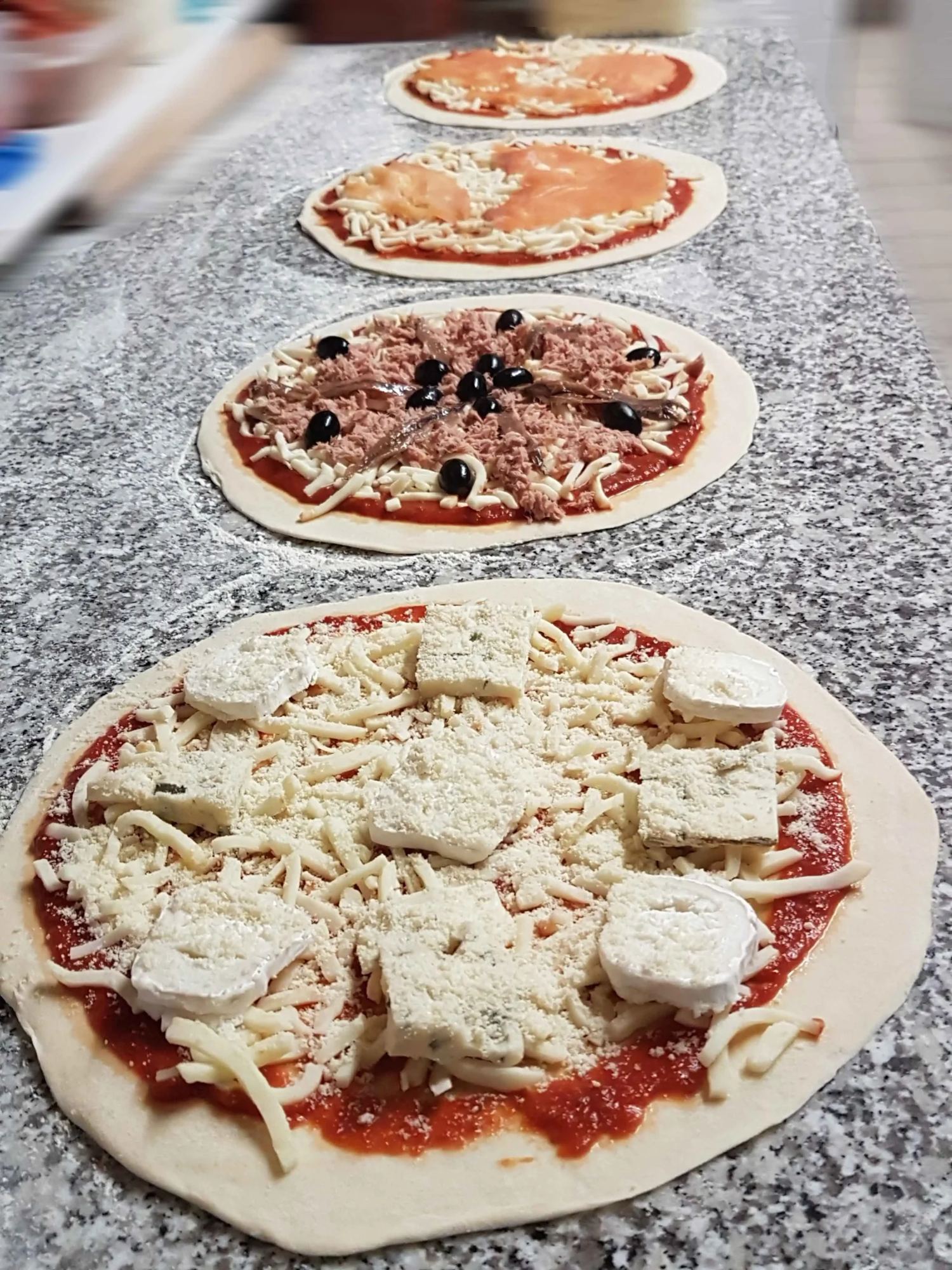 Quatre pizzas crues non cuites, garnies de fromage, de thon, d'olives et de saumon, sont alignées sur un plan de travail en granit saupoudré de farine.