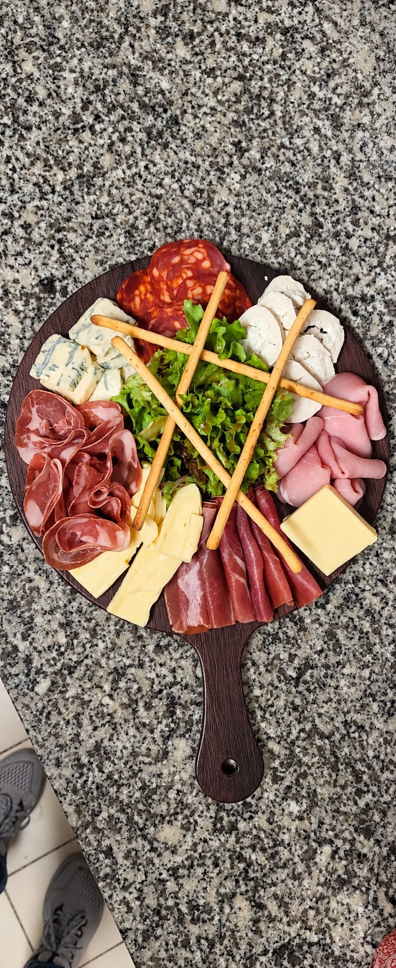 Une planche en bois ronde garnie de charcuteries, de fromages, de gressins et de salade posée sur un plan de travail en granit.