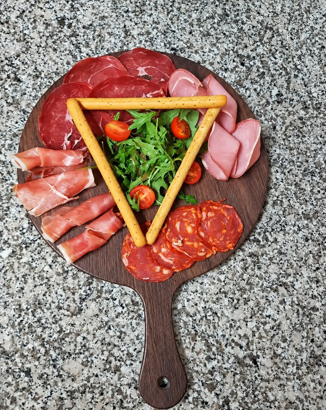 Vue de dessus d'une planche en bois garnie de charcuteries, de roquette, de tomates cerises et de gressins disposés en triangle, posée sur un plan de travail en granit.