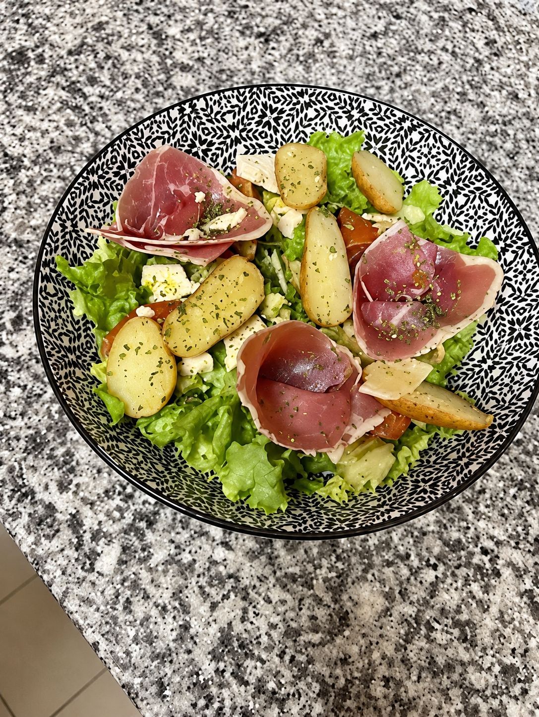Un bol noir et blanc à motifs contenant une salade verte garnie de tranches de jambon cru et de rondelles de pommes de terre, posé sur un plan de travail en granit.
