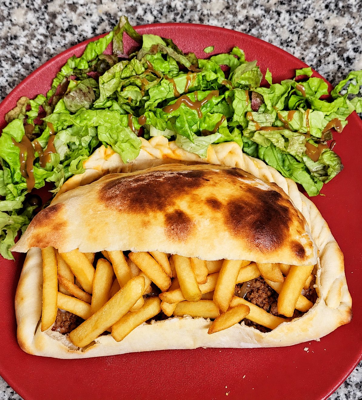 Un sandwich dans un pain pita doré garni de frites et de viande hachée, servi dans une assiette rouge avec de la salade verte.