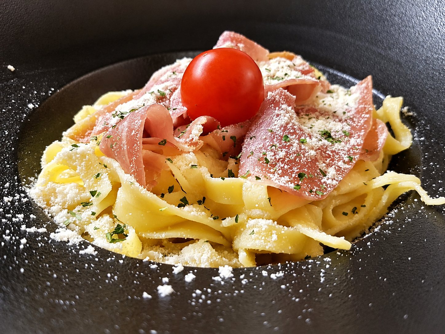 Gros plan sur des tagliatelles fraîches garnies de tranches de jambon cru, de parmesan râpé, de persil et d'une tomate cerise, servies dans une assiette creuse sombre.